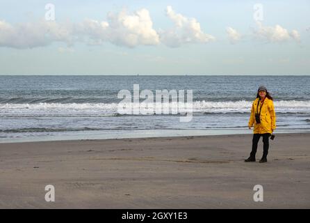 Lady with yellow raincoat on the beach. It's a windy day in autumn. The waves of the North sea are breaking on the shore Stock Photo