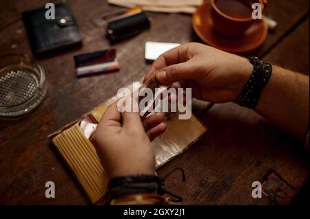 Man rolls a cigarette, wooden table on background. Tobacco smoking culture, specific rich flavor. Male smoker leisures in office Stock Photo