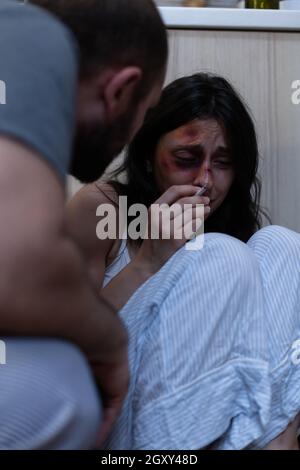 Victim of physical trauma and aggression feeling terrified and scared of violent drunk husband. Alcohol addict threatening scared woman with bruises on face holding cigarette crying Stock Photo