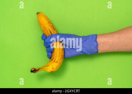 Hand in a latex glove holding a banana on green background with copy space. Grocery stores during COVID-19 lockdown. Stock Photo