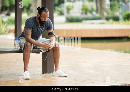Concentrated sporty Indian man with beard sitting on bench in park and listening to music in headphones Stock Photo