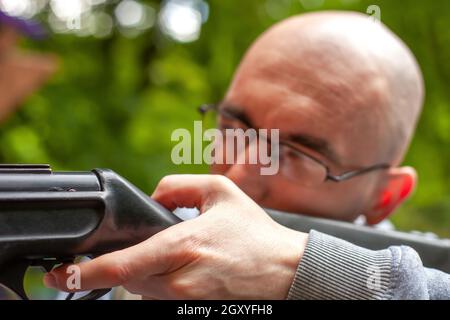 Young man shooting a gun. Man aiming with shotgun on shooting range close up. Hunter in summer green forest closeup, soft selective focus on human han Stock Photo