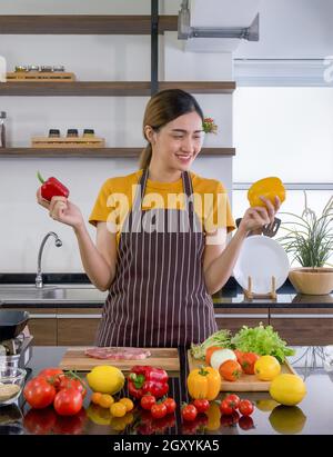 Young housewife stand smiling,  hold red and yellow bell pepper with both hands. Looking at the yellow one on the left. The kitchen counter full of va Stock Photo