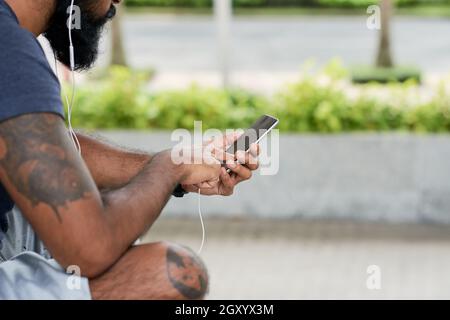 Close-up of bearded man with tattoos listening to audio in earphones and using smartphone outdoors Stock Photo