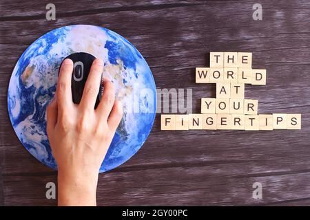 Hand clicking computer mouse on planet earth globe and wooden cube with text THE WORLD AT YOUR FINGERTIPS. Stock Photo