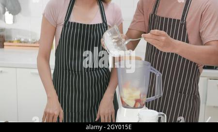 Happy Asian beautiful young family couple husband and wife making fresh apple smoothie in kitchen together at home. The man and woman pour ice into a Stock Photo