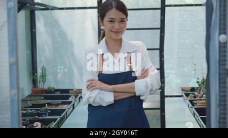 Asian beautiful young gardener woman confident with apron standing posing crossed arms and smiling to camera at greenhouse cactus plant nursery, small Stock Photo