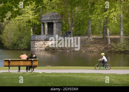 couple sitting on a park bench and a small child riding a bicycle in the city forest, the Deuss temple in the background, Germany, North Stock Photo