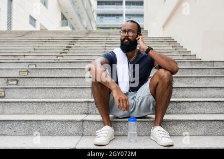 Serious young mixed race man with beard sitting on step and looking into distance while adjusting erphone in ear Stock Photo