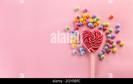 Heart-like striped red lollipop surrounded by sweet colorful candies laying on the pink background. Stock Photo