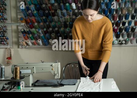 A dressmaker stands at a work table and cuts white fabric along a pattern with scissors. The seamstress in the workshop does what she loves. Stock Photo