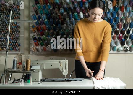 A Caucasian dressmaker stands at a work table and cuts white fabric along a pattern with scissors. The seamstress in the workshop does what she loves. Stock Photo