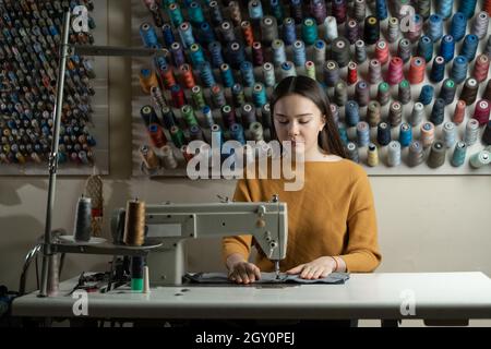 A woman works on a sewing machine in an atelier. She sits at the table and sews cotton clothes. Background from spools of thread of different colors. Stock Photo