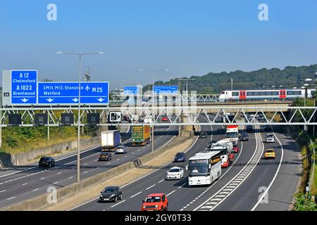 M25 motorway gantry signs railway bridge & Greater Anglia passenger train crossing above road traffic junction 28 for A12 Brentwood Essex England UK Stock Photo