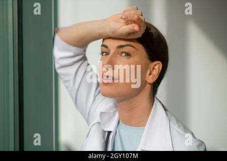 A dark-haired doctor standing near the window and looking thoughtful Stock Photo