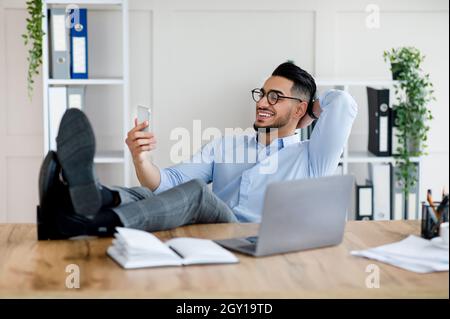 Happy young Arab man in office wear using smartphone, having break, putting feet on table at workplace with laptop Stock Photo