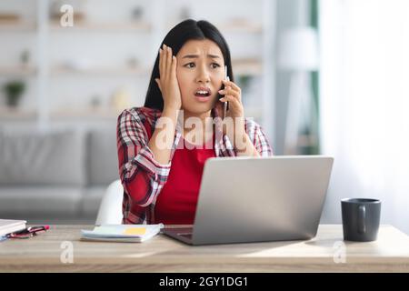 Oh No. Shocked Asian Female Freelancer Talking On Cellphone At Home Office Stock Photo