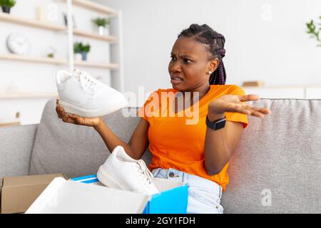 Angry black female opening box from online store and looking at delivered item, upset about received sneaker shoes Stock Photo