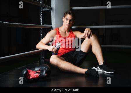 athletic man with sports bottle resting in corner of ring near boxing gloves Stock Photo