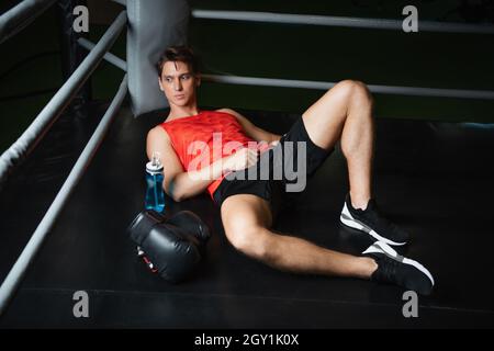 tired sportsman resting in corner of ring near boxing gloves and sports bottle Stock Photo