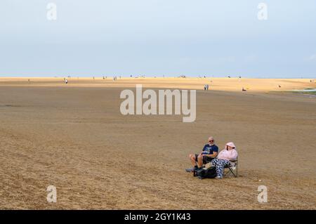 Life is a beach. A couple sitting on a pair of camping chairs on a large sandy beach with plenty of space, Wells Next The Sea, Norfolk, England. Stock Photo