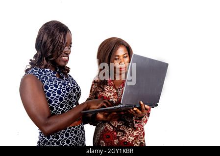 two beautiful African women standing inside holding and working using a laptop while being smiling, isolated on white background Stock Photo