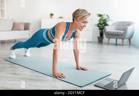 Cheerful woman doing push-ups on mat with laptop Stock Photo