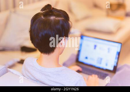 Asian boy with earphone sit on couch working on laptop at home. Stock Photo