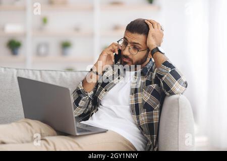 Stressful indian guy with laptop having phone conversation Stock Photo