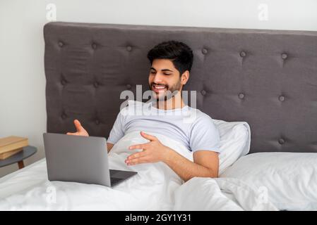 Modern communication concept. Relaxed arab man having video call on laptop computer while sitting in bed Stock Photo