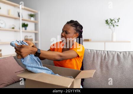 Dissatisfied black female customer opening box from online store, unhappy with received jeans at home Stock Photo