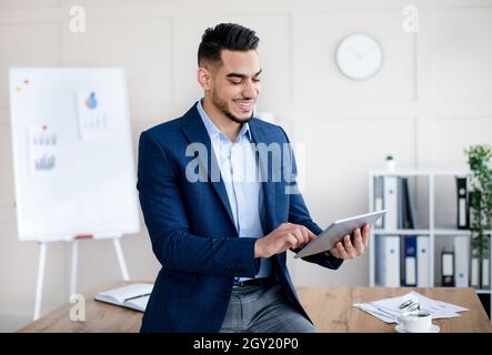 Happy millennial Arab guy using digital tablet, studying or working remotely, having online conference at office Stock Photo