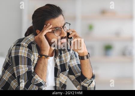 Upset middle-eastern guy having phone conversation, home interior Stock Photo