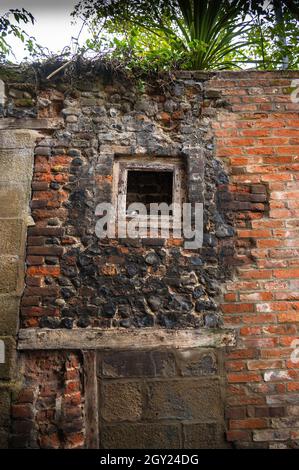 A repaired old flint wall with red brick and large bricks with a small wooden  frame in the wall and weeds growing out the top of wall Stock Photo