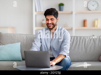 Home office concept. Happy arab guy working on pc laptop, sitting on comfy sofa at home interior, free space Stock Photo
