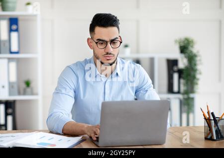 Worried young Arab guy sitting at his desk with laptop computer, making error, concerned about deadline in office Stock Photo