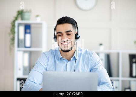 Cheerful Arab man with headphones working online on laptop, having video call at office Stock Photo