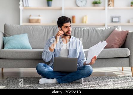 Irritated arab businessman working from home, sitting on floor with laptop, holding documents and having phone call Stock Photo