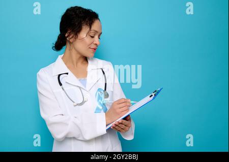 Middle aged mixed race beautiful female doctor wearing a blue satin ribbon, symbolic bow color raising awareness in diabetes day , makingtes on a clip Stock Photo