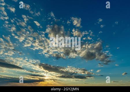 Yellow rays of the setting sun shine in a cloud gap low above the horizon and illuminate a scattering of small clouds high in the sky. Windy weather. Stock Photo