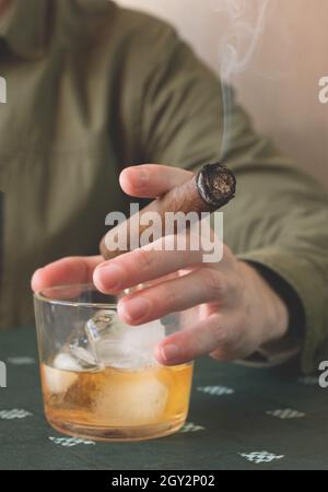 A man holding a Cuban cigar and a glass of whiskey on ice. Close-up of a smoking cigar. Stock Photo
