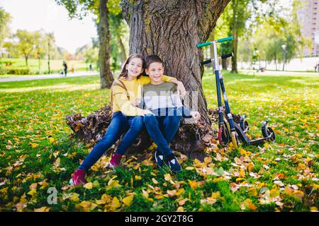 Caucasian boy and girl are hugging under a tree in the park after riding scooters in autumn weather. Portrait of brother and sister enjoying outdoor a Stock Photo