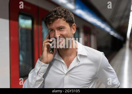 Glad man in white shirt smiling and speaking on cellphone on blurred background of train while commuting to work Stock Photo