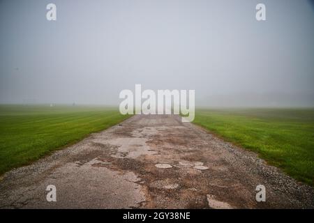 Empty beat-up gravel road in grass field on foggy morning Stock Photo