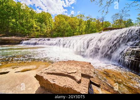 Boulder on coast with large waterfall and lush green forest in background Stock Photo