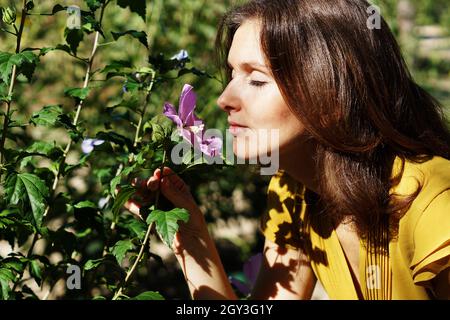 Attractive young Spanish woman smelling fresh purple hibiscus flower in a park Stock Photo