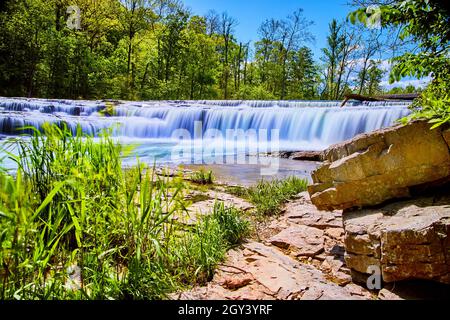 Lush green plants and large rocks with wide waterfall in background Stock Photo