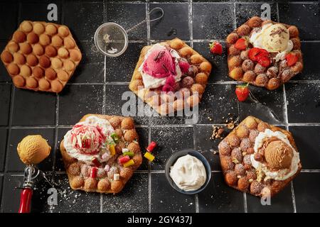 Top down on an assortment of bubble waffles with fresh fruit and nutty ice-cream on a bed of whipped cream drizzled with chocolate shavings and icing Stock Photo