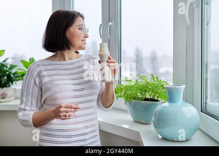 Middle-aged woman drinking milk drink, liquid yogurt in bottle, at home near window Stock Photo