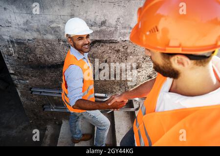 Two men engineers in workwear shaking hands against construction site. Stock Photo